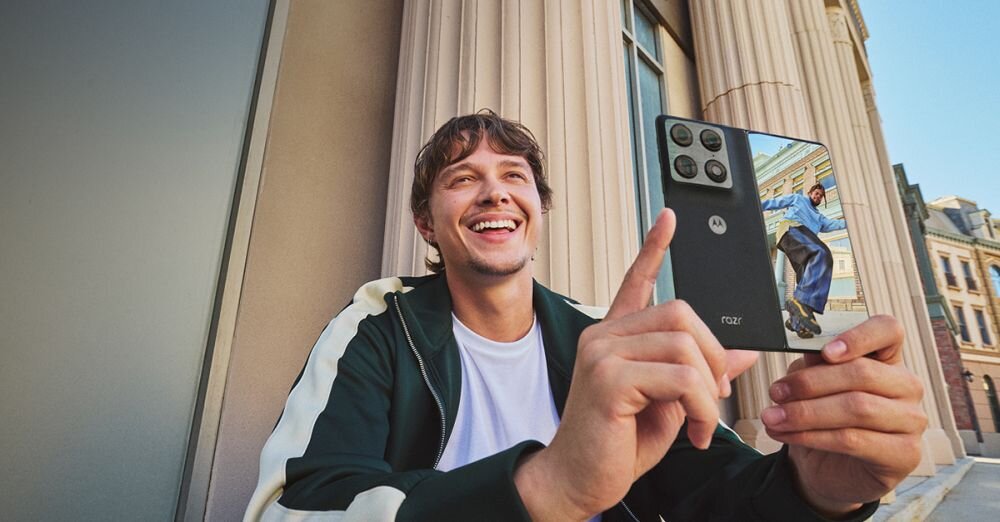 Osoba trzyma smartfon z widocznym logo Motorola i ekranem wyświetlającym zdjęcie mężczyzny w niebieskim stroju. W tle znajdują się kolumny budynku i fragmenty architektury miejskiej.