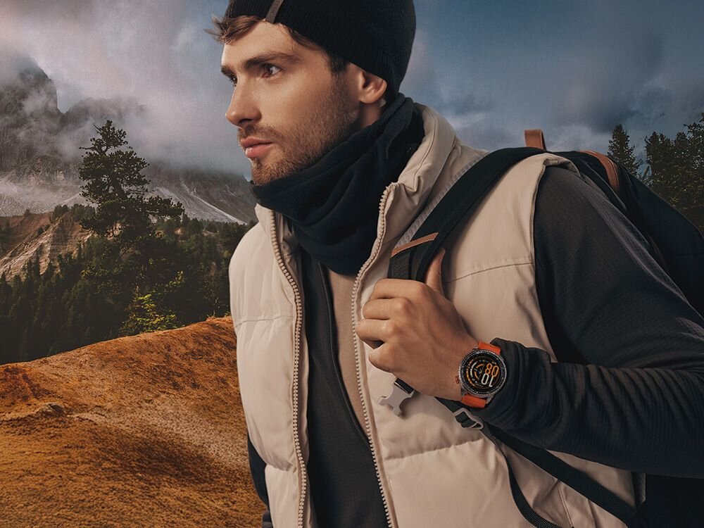 Osoba ubrana w zimową odzież i plecak stoi na tle górskiego krajobrazu. Na nadgarstku ma smartwatch z widocznym okrągłym wyświetlaczem.
