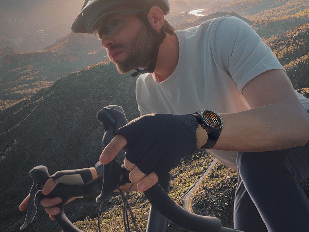Osoba w kasku rowerowym i rękawiczkach trzyma kierownicę roweru, na nadgarstku ma smartwatch z okrągłym wyświetlaczem. W tle widoczny jest górski krajobraz.