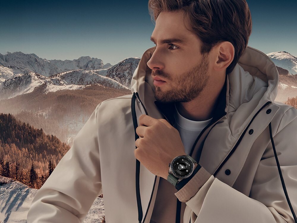 Osoba w jasnej kurtce patrzy w dal, na nadgarstku ma smartwatch. W tle widać ośnieżone góry i lasy.