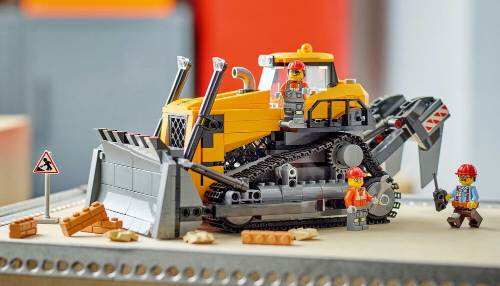 Na obrazie znajduje się żółty buldożer z klocków LEGO, otoczony przez trzy figurki w kaskach. Obok buldożera widoczne są cegły i znak ostrzegawczy z symbolem prac drogowych.
