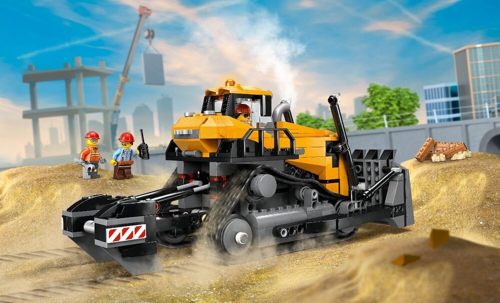 Na obrazie znajduje się żółty buldożer z klocków LEGO na placu budowy, otoczony przez dwie figurki pracowników w kaskach. W tle widoczne są elementy konstrukcyjne i zarys miasta.
