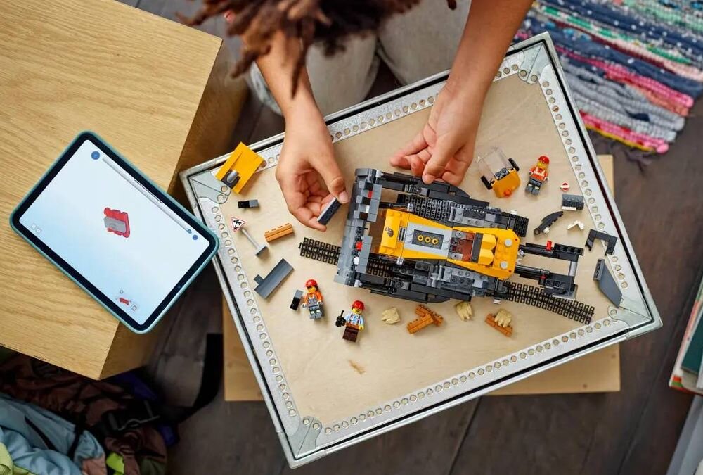 Osoba składa model żółtego buldożera z klocków LEGO na stole, wokół znajdują się różne elementy zestawu. Obok leży tablet z wyświetlonym schematem budowy.