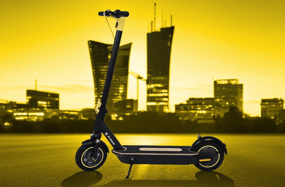 Czarna hulajnoga elektryczna z napisem \'XRIDER\' stoi na tle miejskiego krajobrazu z żółtym niebem. W tle widoczne są nowoczesne budynki i wieżowce.
