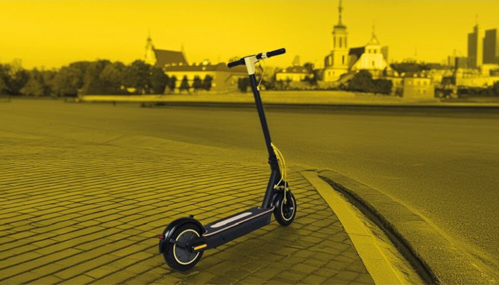 Czarna hulajnoga elektryczna stoi na chodniku z widokiem na miasto w tle. Obraz ma żółty filtr, a w tle widoczne są budynki i drzewa.