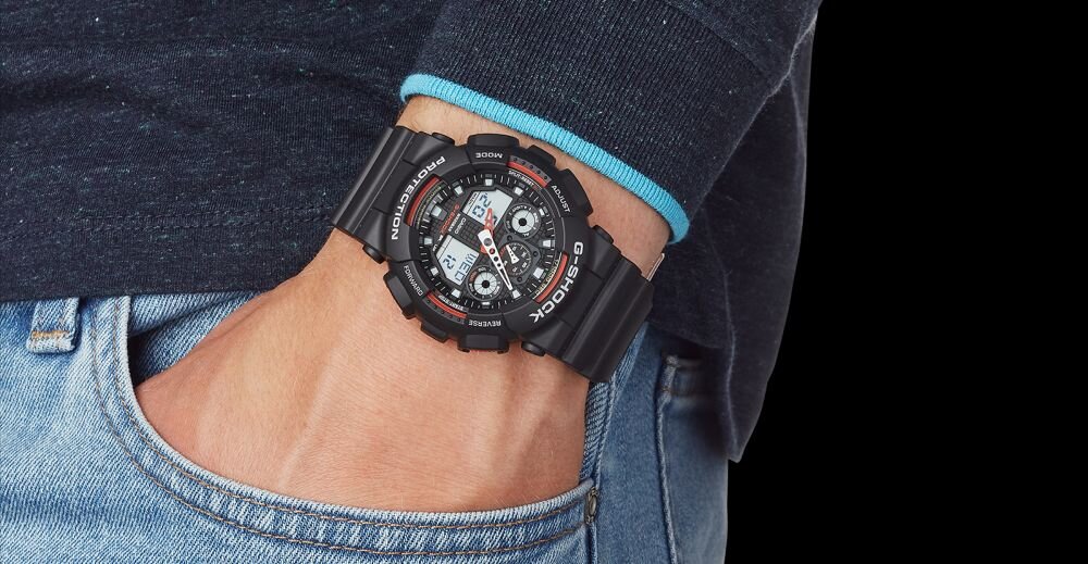 Osoba nosi czarny zegarek sportowy z dużą tarczą i cyfrowymi wyświetlaczami, na którym widoczne są napisy \'G-SHOCK\' i \'PROTECTION\'. Zegarek ma gumowy pasek i jest założony na nadgarstek osoby ubranej w ciemny sweter i dżinsy.