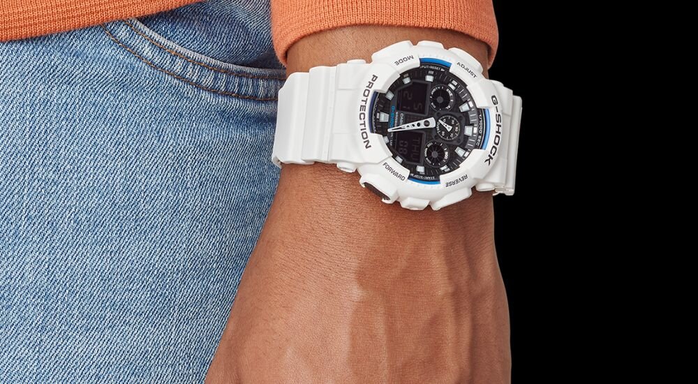 Osoba nosi biały zegarek sportowy z dużą tarczą i cyfrowym wyświetlaczem. Na zegarku widoczne są napisy \'PROTECTION\' i \'G-SHOCK\'.