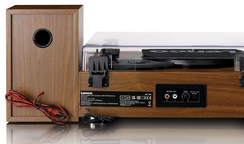 Na obrazie widoczny jest gramofon z drewnianą obudową oraz kolumna głośnikowa. Na gramofonie znajduje się logo \'Lenco\' oraz różne złącza i napisy techniczne.
