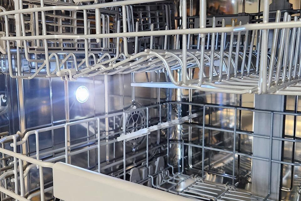 Tub Light czyli oświetlenie wnętrza zmywarki Wnętrze zmywarki z widocznymi metalowymi koszami na naczynia i oświetleniem wewnętrznym. W tle widać błyszczącą, metalową powierzchnię.