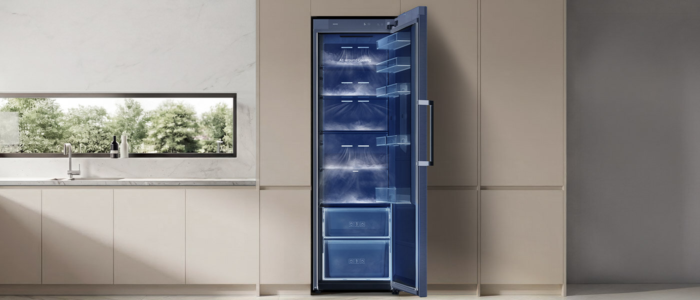 Chłodziarka z technologią All Around Cooling Otwarta lodówka w kolorze szczotkowanej stali nierdzewnej z widocznymi półkami i szufladami wewnątrz. W tle znajduje się nowoczesna kuchnia z jasnymi szafkami i dużym oknem.