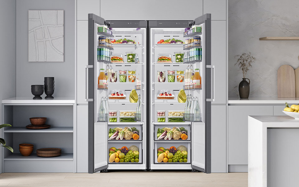 Przekładane drzwi Obraz przedstawia otwartą lodówkę z widocznymi półkami wypełnionymi różnorodnymi produktami spożywczymi, takimi jak owoce, warzywa, napoje i słoiki. Lodówka ma nowoczesny design i jest umieszczona w kuchni z szarymi szafkami i dekoracjami.