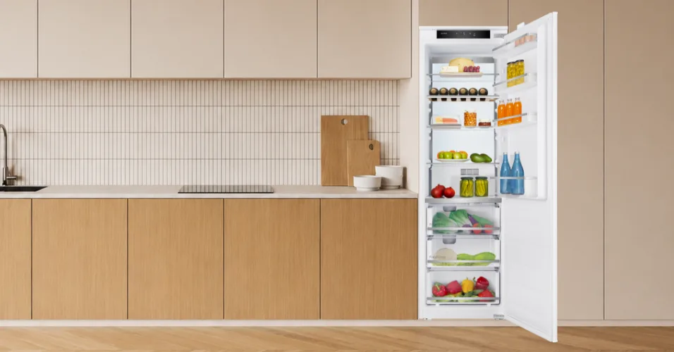 Wnętrze kuchni z otwartą lodówką w zabudowie, wypełnioną różnorodnymi produktami spożywczymi, takimi jak owoce, warzywa, napoje i słoiki. Obok lodówki znajduje się blat z drewnianymi frontami i zlewem.