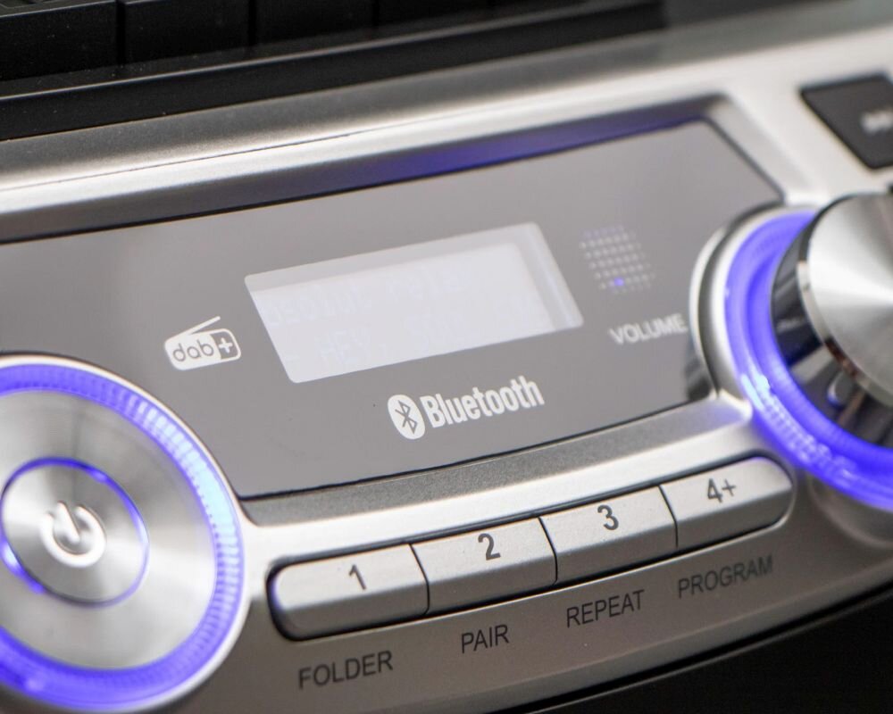 Zbliżenie na panel sterowania radioodtwarzacza z widocznymi przyciskami i pokrętłami. Na panelu znajdują się napisy: \'Bluetooth\', \'FOLDER\', \'PAIR\', \'REPEAT\', \'PROGRAM\', oraz symbole DAB+ i Bluetooth.