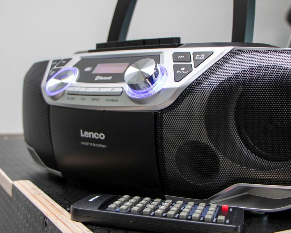 Radioodtwarzacz czarno-srebrny z widocznym logo \'Lenco\' i napisem \'CASSETTE RECORDER\'. Na panelu znajdują się przyciski i pokrętło, a przed urządzeniem leży pilot z przyciskami.