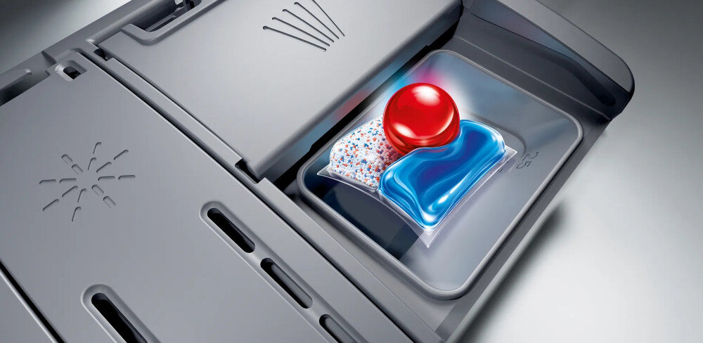 Obraz przedstawia wnętrze zmywarki z otwartym pojemnikiem na detergent, w którym znajduje się kolorowa kapsułka do mycia naczyń. Pojemnik ma szary kolor i widoczne są na nim delikatne wzory oraz linie.