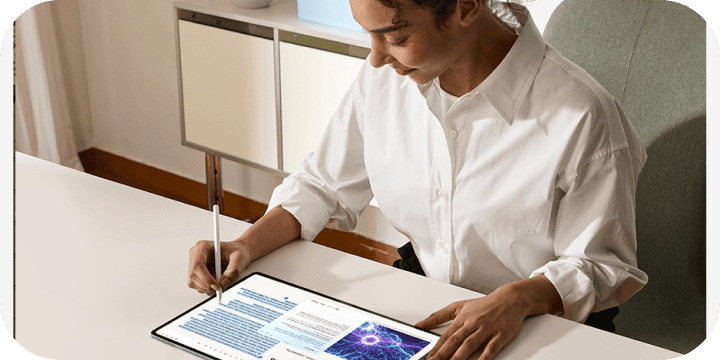 Osoba korzystająca z tabletu Samsung Galaxy i rysika S Pen przy biurku Osoba siedzi przy biurku i używa rysika do pisania na dużym tablecie, na którym wyświetlany jest tekst i obraz. Tablet leży na białym blacie, a w tle widać fragment szafki i krzesła.
