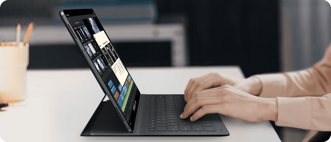 Tablet Galaxy z podłączoną klawiaturą umożliwiającą pracę jak na laptopie. Osoba używa tabletu z klawiaturą, na ekranie widoczne są różne aplikacje i dokumenty. Tablet jest ustawiony na biurku w jasnym pomieszczeniu.