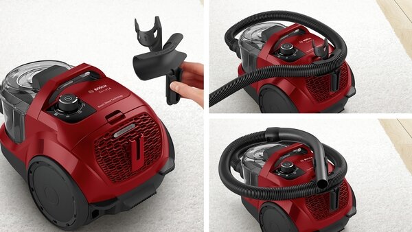 Na obrazie widoczny jest czerwony odkurzacz z logo BOSCH, z przezroczystym pojemnikiem na kurz i czarnym wężem. Obok odkurzacza znajduje się ręka trzymająca czarną plastikową część.