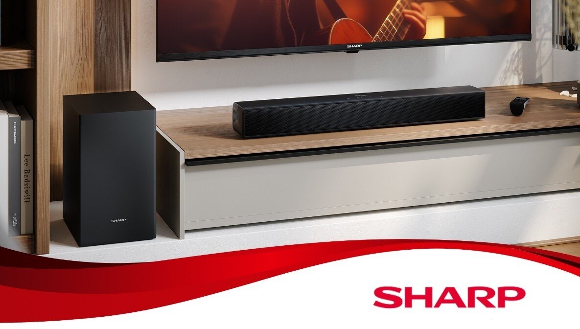 Na obrazie znajduje się czarny soundbar umieszczony na drewnianej półce pod telewizorem. W dolnej części obrazu widoczne jest czerwone logo z napisem \'SHARP\'.