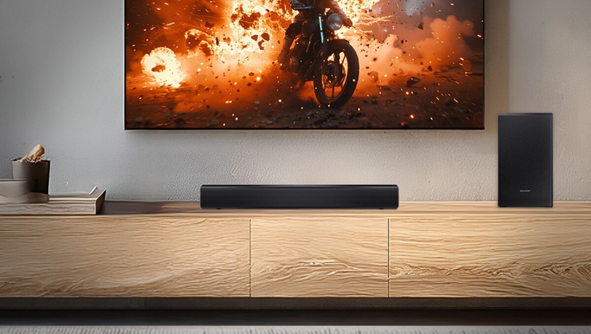 Na drewnianej szafce stoi czarny soundbar oraz subwoofer. Na ścianie wisi telewizor z dynamiczną sceną eksplozji i motocykla.