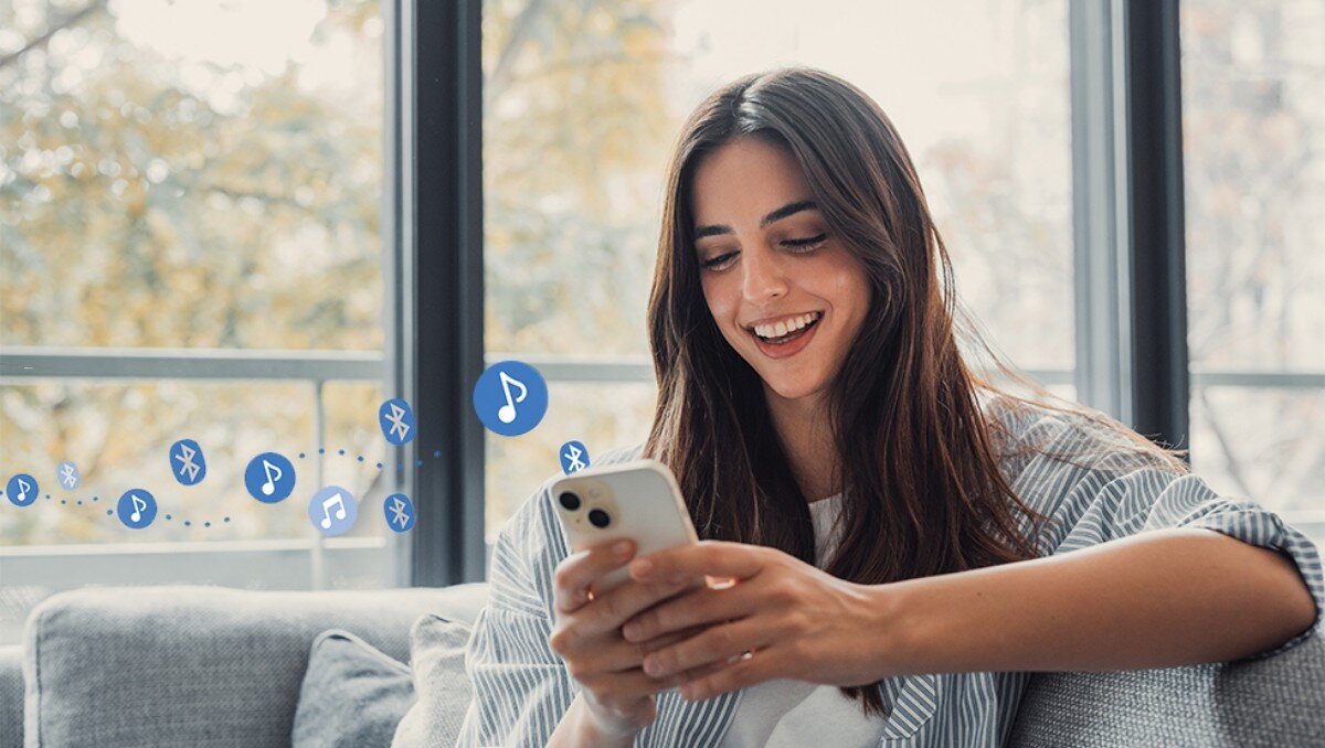 Osoba siedzi na kanapie, trzymając w ręku smartfon. W tle widać ikony Bluetooth i nut, sugerujące przesyłanie muzyki.