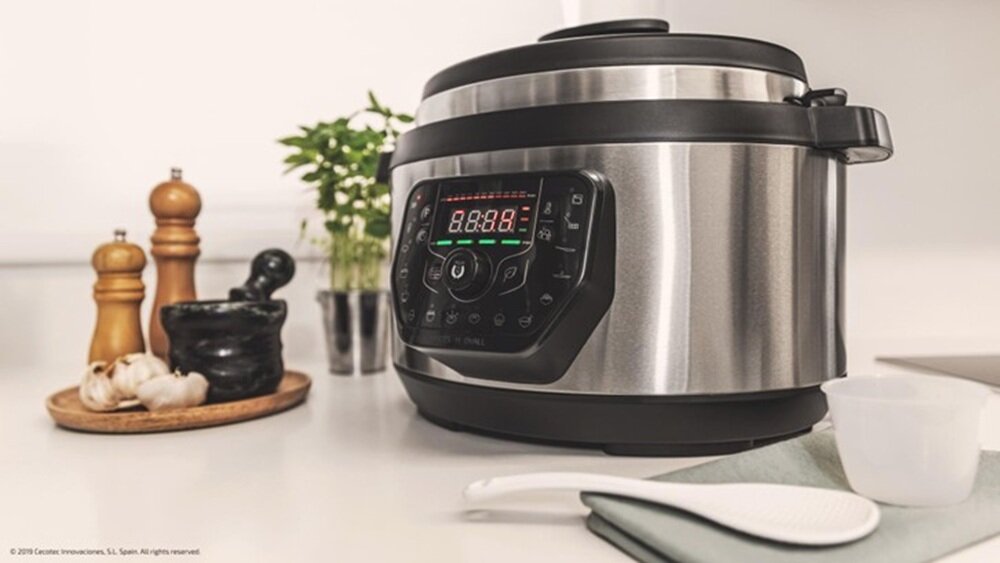 Na blacie kuchennym stoi multicooker ze stalowym korpusem i czarnym panelem sterowania z wyświetlaczem LED. Obok znajdują się drewniane młynki do przypraw, czosnek oraz zielona roślina.
