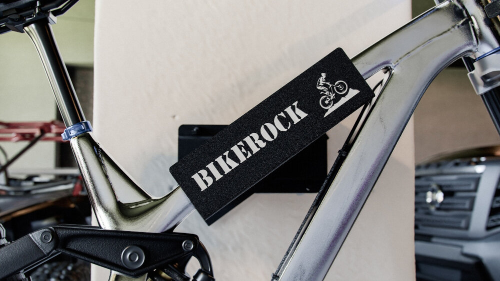 Na obrazie widoczny jest fragment roweru zamocowanego na wieszaku z napisem \'BIKEROCK\' oraz grafiką rowerzysty. Wieszak jest przymocowany do ściany.