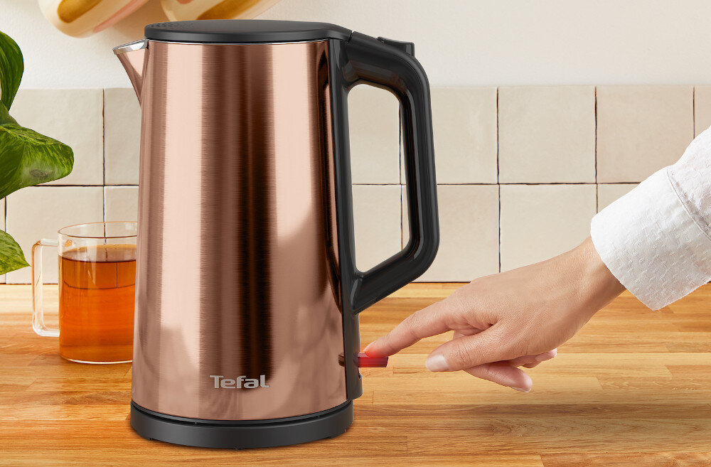 Na drewnianym blacie stoi metaliczny czajnik elektryczny z czarnym uchwytem i napisem \'Tefal\'. Obok czajnika znajduje się szklanka z herbatą, a ręka naciska przycisk na podstawie czajnika.