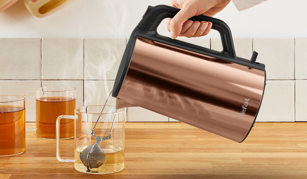 Osoba nalewa gorącą wodę z metalicznego czajnika do szklanki z herbatą. Na czajniku widoczne jest logo \'Tefal\'.