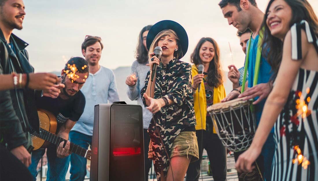 Grupa ludzi bawi się na zewnątrz, w centrum widoczny jest głośnik Power Audio SAMSUNG MX-T70. Osoba trzyma mikrofon, a inni grają na instrumentach i trzymają zimne ognie.