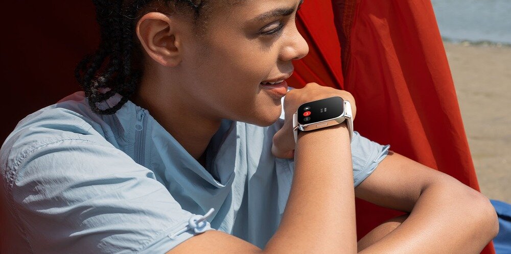 Osoba siedzi na zewnątrz, opierając się o czerwoną tkaninę, i patrzy na smartwatch na swoim nadgarstku. Smartwatch ma czarny ekran z widocznymi ikonami aplikacji.