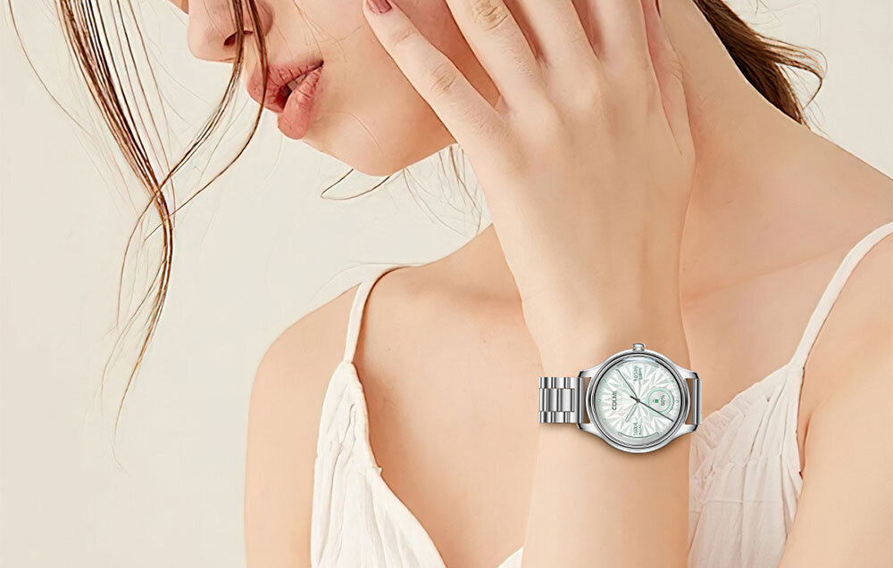 Osoba w białej bluzce prezentuje na nadgarstku złoty smartwatch z metalową bransoletą i okrągłą tarczą. Tło jest jednolite i jasne.