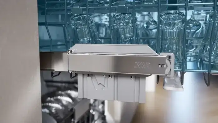 Wnętrze zmywarki z widocznymi szklankami ustawionymi w górnym koszu. Na pierwszym planie znajduje się metalowy element z logo producenta, a w tle widać dolny kosz z naczyniami.