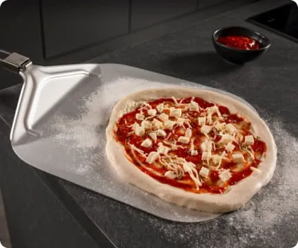 Na blacie kuchennym leży surowa pizza na metalowej łopacie, obok znajduje się miseczka z sosem pomidorowym.