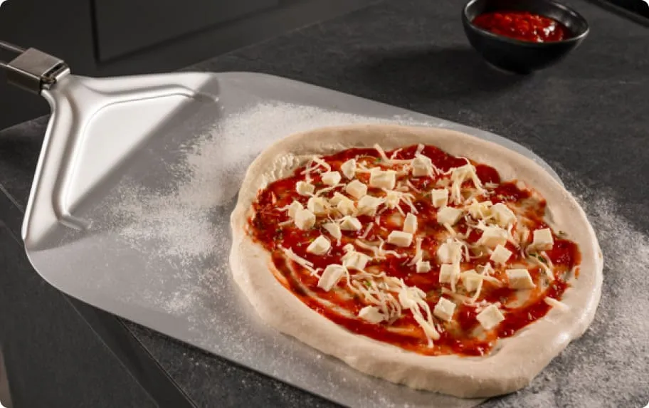 Na metalowej łopacie do pizzy znajduje się surowa pizza z sosem pomidorowym, posypana kawałkami sera. W tle widoczna jest miseczka z czerwonym sosem.