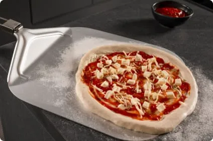 Na obrazie widoczna jest surowa pizza na metalowej łopacie, obok znajduje się miseczka z sosem pomidorowym. Powierzchnia jest posypana mąką.