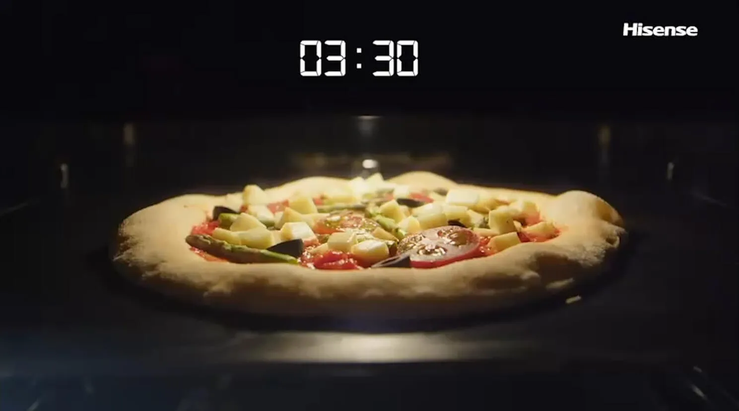 W piekarniku znajduje się pizza z różnymi składnikami, a na wyświetlaczu widoczny jest czas 03:30. W prawym górnym rogu znajduje się logo \'Hisense\'.