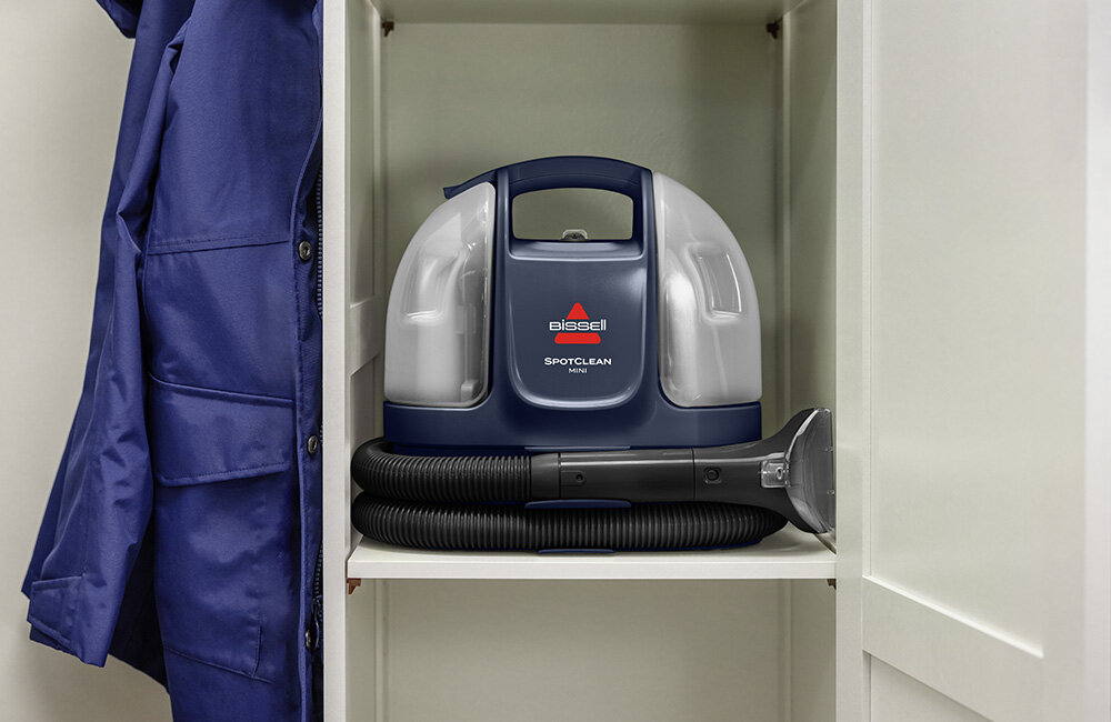 Odkurzacz piorący BISSELL SpotClean Mini Pro 4149N umieszczony na półce w szafie. Obok wisi niebieska kurtka.