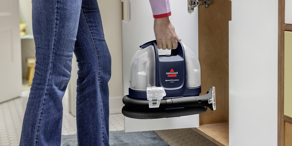 Osoba trzyma odkurzacz piorący z widocznym logo \'BISSELL\' i napisem \'SpotClean\'. Odkurzacz jest wyciągany z szafki w pomieszczeniu z jasną podłogą.