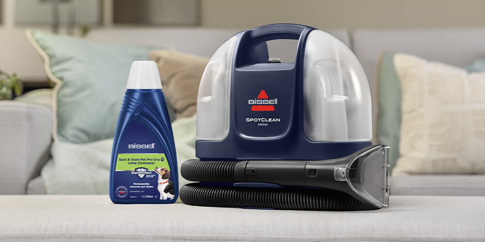 Na obrazie znajduje się odkurzacz piorący z napisem \'BISSELL SpotClean Mini\' oraz butelka środka czyszczącego z logo \'Bissell\'. W tle widoczna jest kanapa z poduszkami.