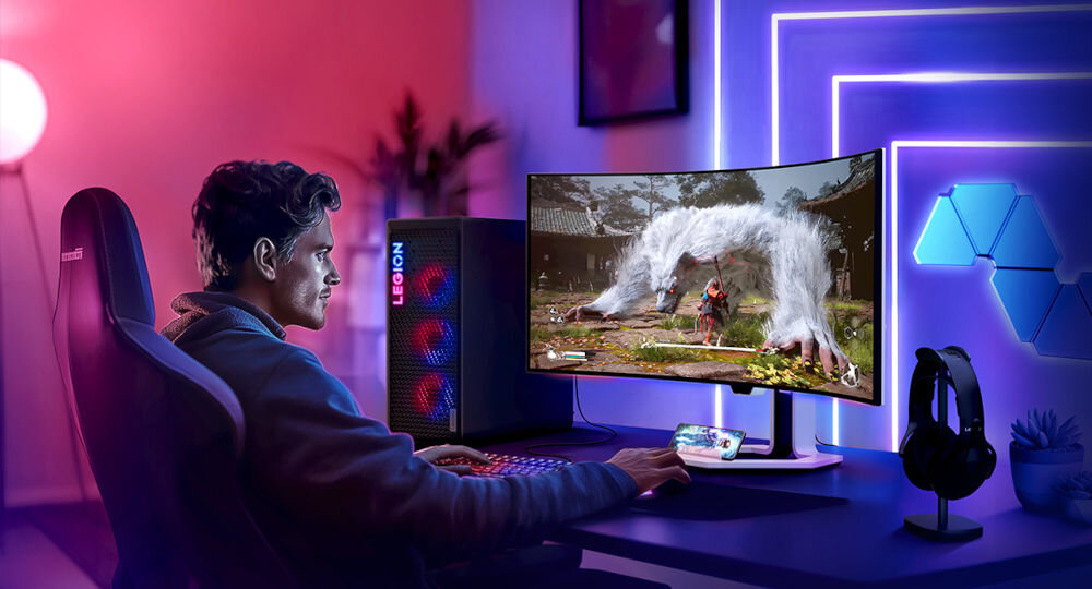 Osoba siedzi przy biurku, grając na zakrzywionym monitorze, na którym wyświetlana jest dynamiczna scena z gry. Obok monitora znajduje się komputer z widocznym logo \'LEGION\'.