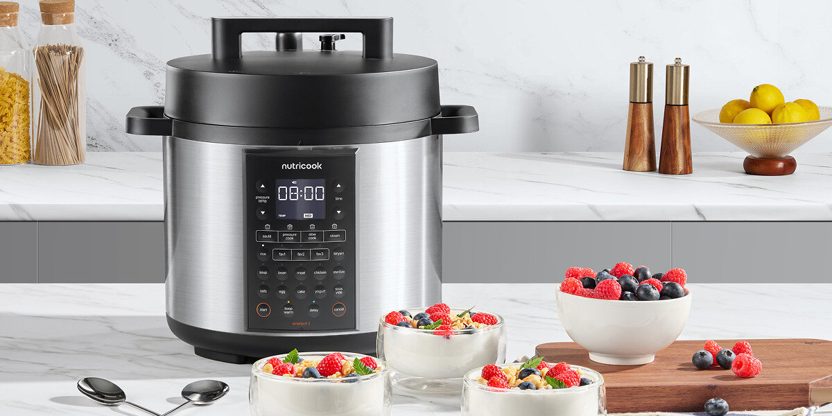 Na blacie kuchennym stoi multicooker Nutricook z wyświetlaczem cyfrowym i przyciskami. Obok znajdują się miski z jogurtem i owocami oraz przyprawy i owoce w tle.