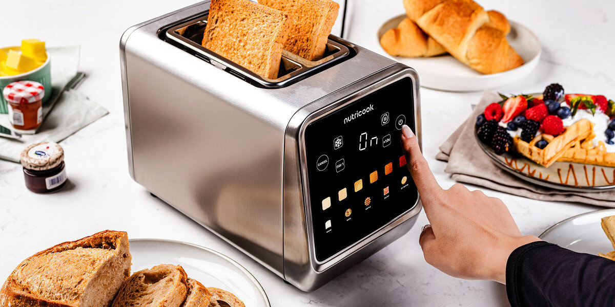 Na obrazie widoczny jest toster Nutricook z dwoma kromkami chleba, a obok niego różne pieczywo i owoce. Na panelu tostera znajdują się przyciski i wyświetlacz z napisem \'On\'.