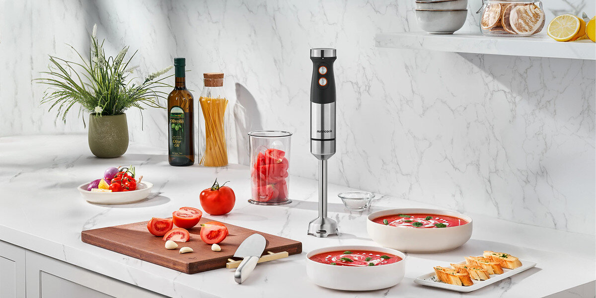 Na blacie kuchennym znajduje się blender ręczny, obok którego leżą pokrojone pomidory i czosnek na desce do krojenia. W tle widoczne są butelki z oliwą, makaron w słoiku oraz miski z zupą i pieczywem.