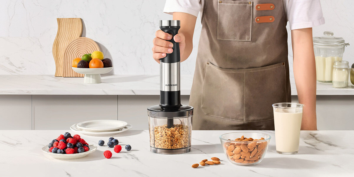 Osoba trzyma blender z logo \'nutricook\', który stoi na blacie obok miski z orzechami, szklanki mleka i talerza z owocami. W tle widać dekoracje i pojemniki kuchenne.