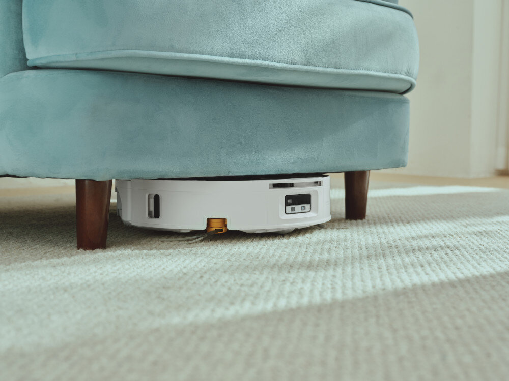 Biały robot sprzątający znajduje się pod niebieską sofą na jasnym dywanie. Robot ma prostokątny panel z przyciskami na górze.