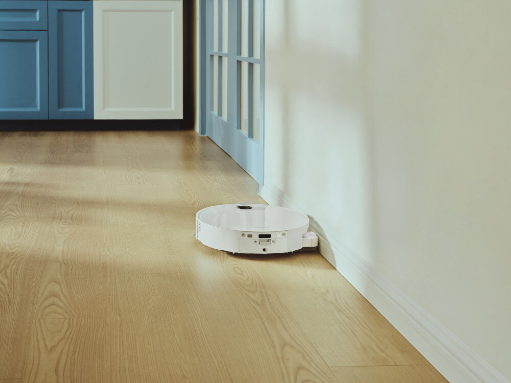 Biały robot sprzątający porusza się wzdłuż ściany na drewnianej podłodze. W tle widoczne są niebieskie drzwi i szafki.