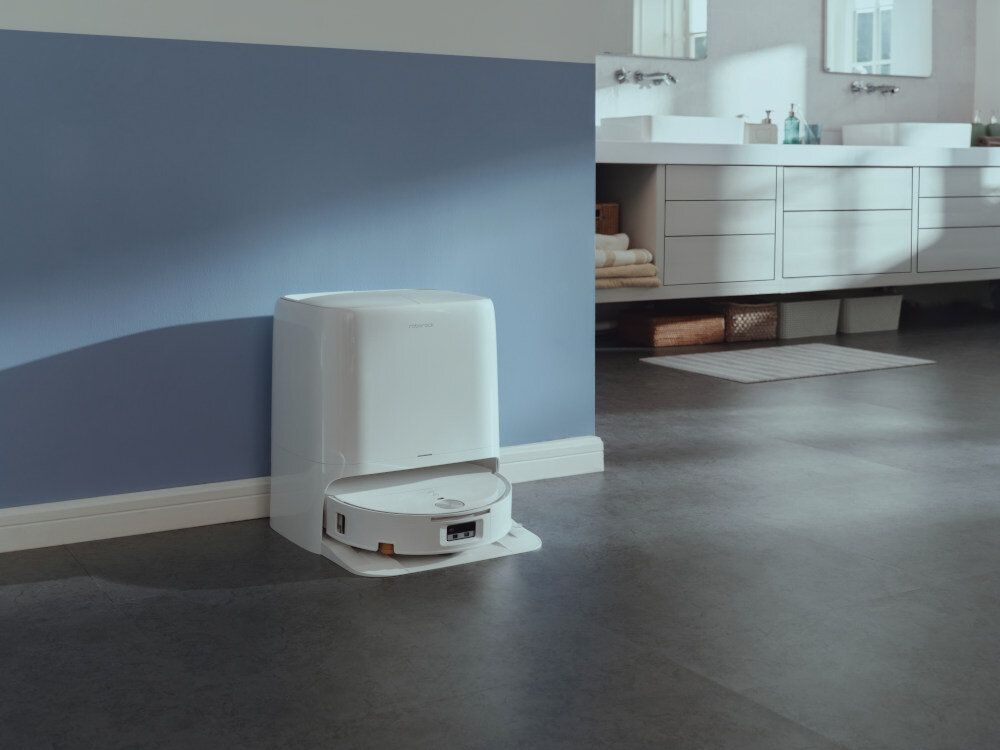 Na obrazie znajduje się biały robot sprzątający w stacji dokującej, ustawiony przy ścianie w pomieszczeniu z niebieską ścianą i ciemną podłogą. W tle widoczna jest łazienka z umywalkami i szafkami.