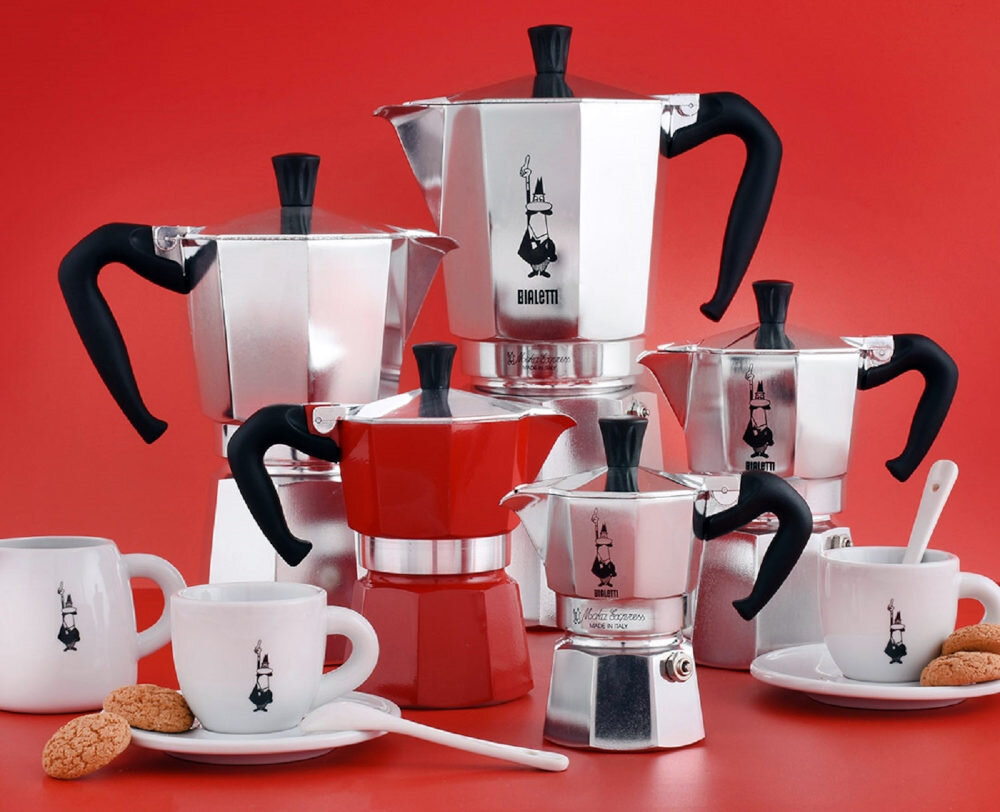 Na czerwonym tle znajduje się kilka kawiarek Bialetti, w tym jedna czerwona, oraz filiżanki z logo Bialetti. Obok filiżanek leżą ciastka i łyżeczki.