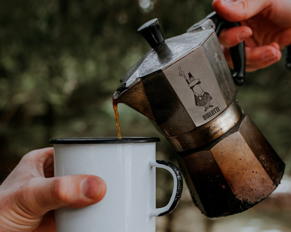 Osoba nalewa kawę z metalowej kawiarki do białego kubka emaliowanego. Na kawiarce widoczne jest logo BIALETTI.